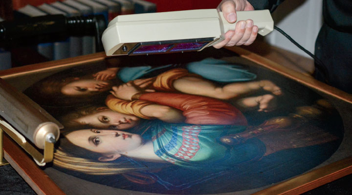 A person holds a UV scanner over a framed painting of two figures