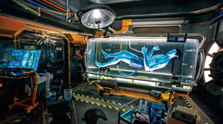 A blue humanoid figure floats inside a glowing pod in a futuristic laboratory setting