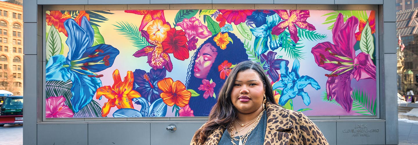 Photo of artist standing in front of their mural in a city